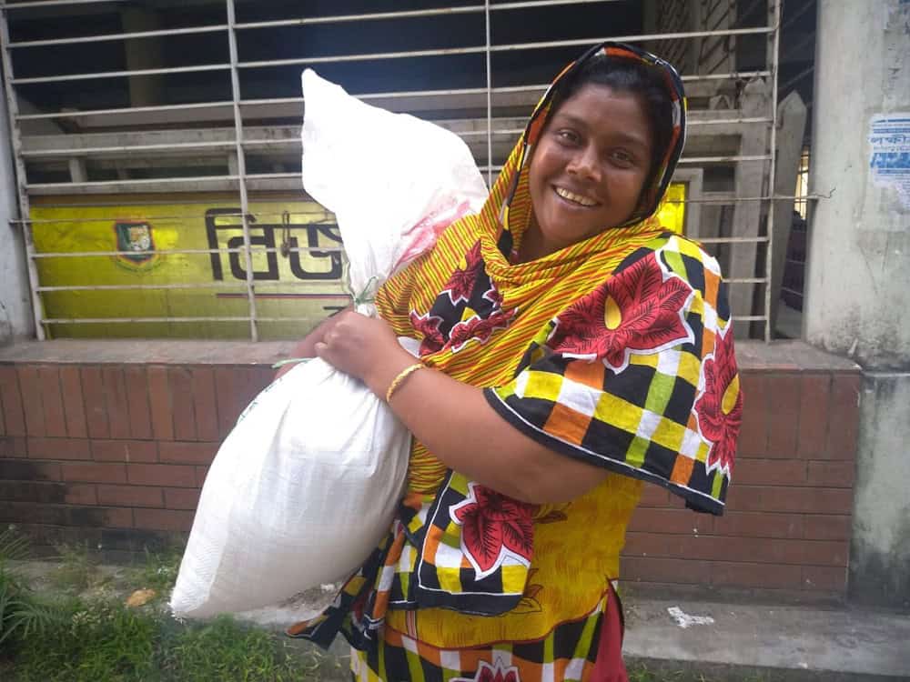 স্মিত হাসি সহ একজন নারী সুবিধাবঞ্চিত পরিবারের পাশে দাঁড়িয়ে. Channel i News - চ্যানেল আই নিউজ