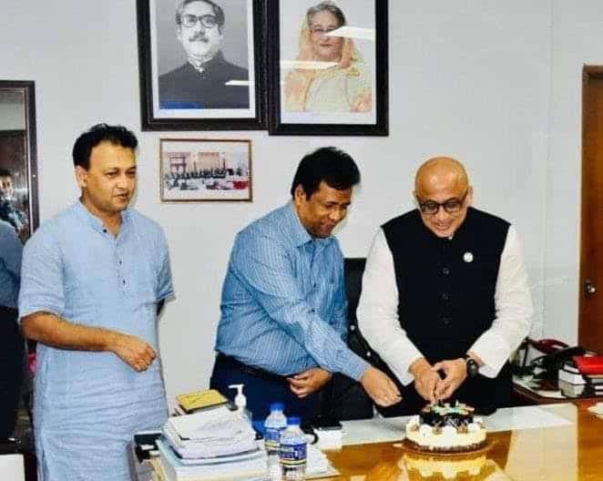 তিনজন ব্যক্তি একটি টেবিলের উপর কেক কাটছেন, পেছনে ছবি ও বই দেখানো. Channel i News - চ্যানেল আই নিউজ