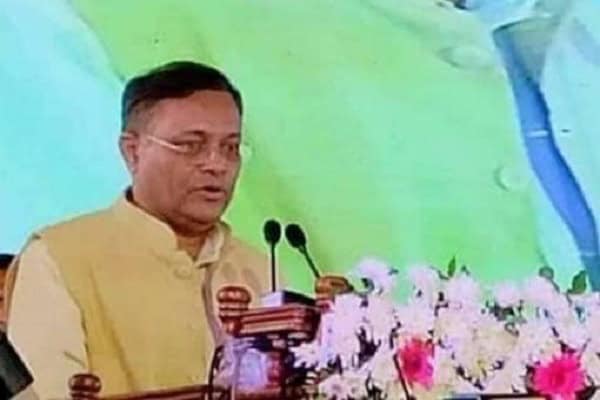 প্রধানমন্ত্রী বক্তৃতা দিচ্ছেন, পেছনে ফুলের সাজানো ব্যাকড্রপ. Channel i News - চ্যানেল আই নিউজ