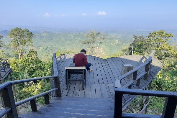 Person sitting alone on a wooden viewing platform overlooking lush green hills. Channel i News - চ্যানেল আই নিউজ