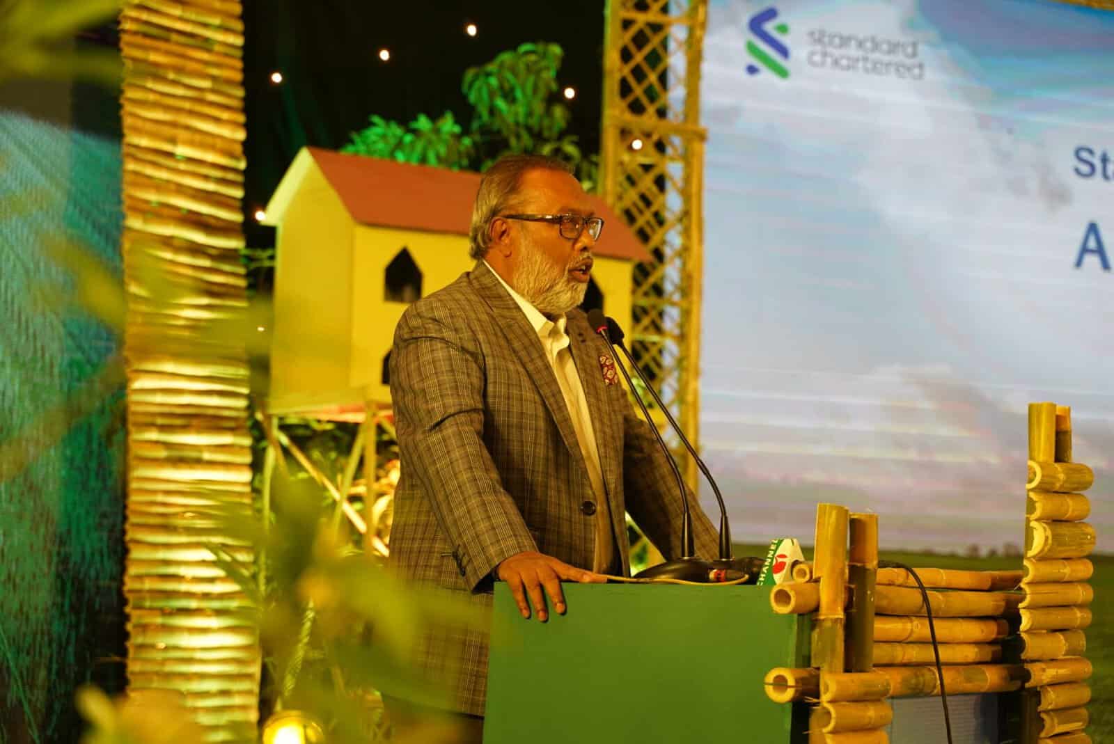 প্রধান অতিথি বক্তব্য দিচ্ছেন একজন পুরুষ, পেছনে স্টেজ ও স্ক্রিন দেখা যাচ্ছে. Channel i News - চ্যানেল আই নিউজ