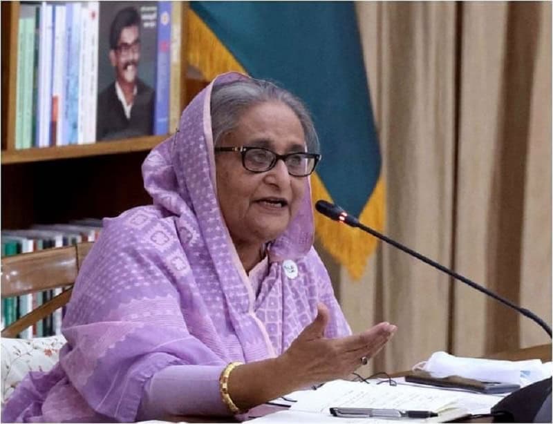 প্রধানমন্ত্রী শেখ হাসিনা ভাষণ দিচ্ছেন, পেছনে বাংলাদেশের পতাকা ও বইয়ের শেলফ. Channel i News - চ্যানেল আই নিউজ