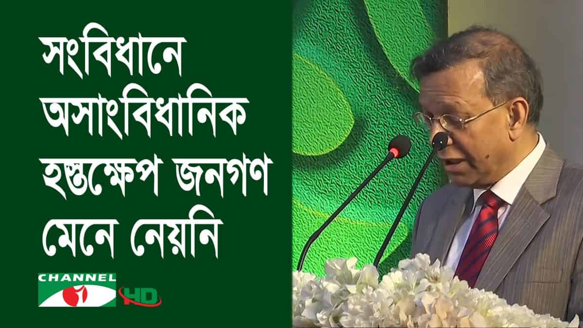আইনমন্ত্রী বক্তৃতা দিচ্ছেন, পেছনে সবুজ ব্যাকড্রপ ও ফুলের তোড়া. Channel i News - চ্যানেল আই নিউজ