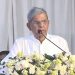 বয়সী ব্যক্তি বক্তৃতা দিচ্ছেন, পেছনে সাদা পর্দা ও ফুলের সাজ. Channel i News - চ্যানেল আই নিউজ