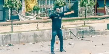 রাস্তার মধ্যে একজন ব্যক্তি বক্তৃতা দিচ্ছেন, চারপাশে কিছু গাছ ও দোকান দেখা যাচ্ছে. Channel i News - চ্যানেল আই নিউজ